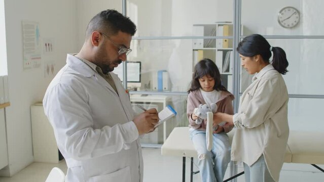 Medium slowmo of Middle Eastern male pediatrician filling in patient prescription paper while Hispanic little girl with soft toy and mom waiting in background