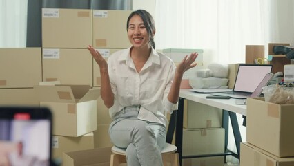 Youth Asia lady owner small business online shop sit front of phone look at camera live streaming talk to customer order promotion with full carton box at home. Shipping and delivery concept.