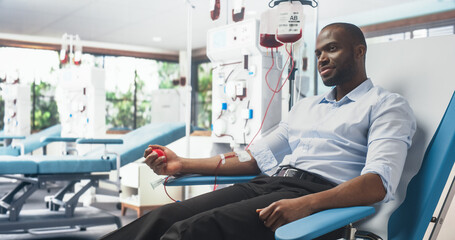 Black Man Donating Blood For People In Need In Bright Hospital. African Male Donor Squeezing Heart-Shaped Red Ball To Pump Blood Through The Tubing Into The Bag. Donation for Children Battling Cancer.