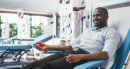 Black Businessman Donating Blood For People In Need In Bright Hospital. Male Donor Squeezing Heart-Shaped Red Ball To Pump Blood Through The Tubing Into The Bag. Donation For Patients Battling Cancer.