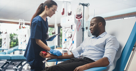 Black Businessman Donating Blood For People In Need In Bright Hospital. Female Nurse With Tablet Computer Coming In To Check Progress And Well-Being Of Donor. Donation For Heart Surgery Patients. - obrazy, fototapety, plakaty