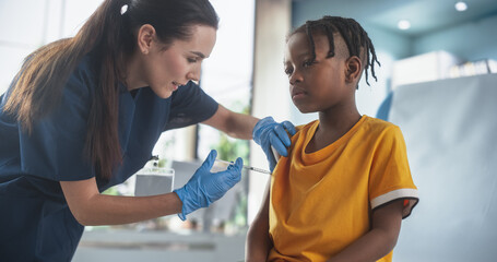 Young African American Boy Sitting In The Chair In Bright Hospital And Getting His Flu Vaccine. Caucasian Female Nurse Is Performing Injection. Professional Woman Talks To Worried Kid, Calms Him Down. - obrazy, fototapety, plakaty