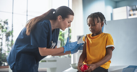Young African American Boy Sitting In The Chair In Bright Hospital And Getting Polio Vaccine. Caucasian Female Nurse Is Performing Injection. Professional Woman Talks To Worried Kid, Calms Him Down. - obrazy, fototapety, plakaty