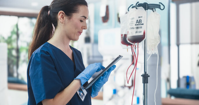 Female Nurse Holding Tablet Computer And Checking The Blood Bag In Donation Center. Professional Caucasian Woman Observing Donation Process In Bright Hospital. Healthcare And Charity Concept.
