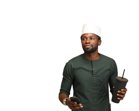 png of Influential businessman with smartphone and portable cup. A handsome executive looking away while standing against a brown studio wall. He is wearing African green attire with white native cap
