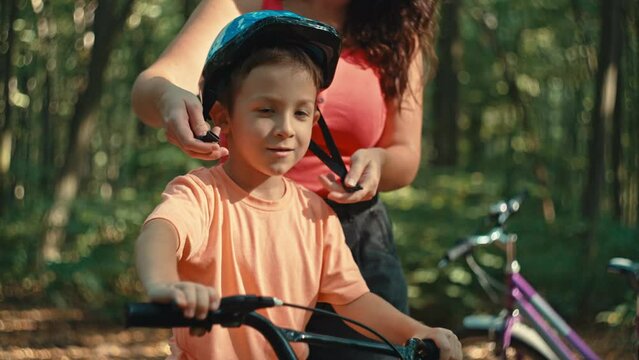 Mom Puts Her Son On A Protective Bike Helmet In The Park While Riding. Protection And Safety When Children Are Cycling In The Forest. High Quality 4k Footage