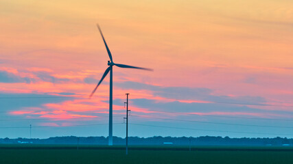 Gorgeous pink and blue sunset clouds behind spinning wind turbine in green field