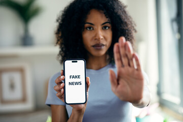 Serious woman holding the mobile phone with the message fake news while doing stop sign with hand...