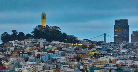 Coit Tower lit up at dusk with buildings below hill and Oakland Bay Bridge in distance aerial