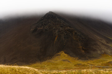 Rocky black volcanic mountain with peak visible through low-altitude clouds after heavy rain in Snaefellsnes peninsula in Iceland.