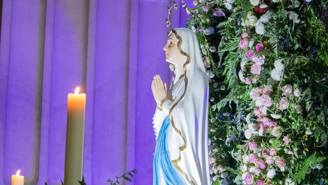 Image of Our Lady of Lourdes seen from the side with colorful flower arrangements in the background inside a church during a solemn celebration