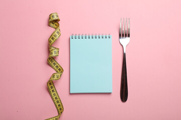 Fork with measuring tape and notebook on pink background. Weight loss, diet concept - obrazy, fototapety, plakaty