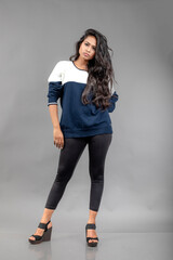 Indian young female model in casual winterwear against grey background. Long black haired model wearing black leggings, blue and white sweatshirt.