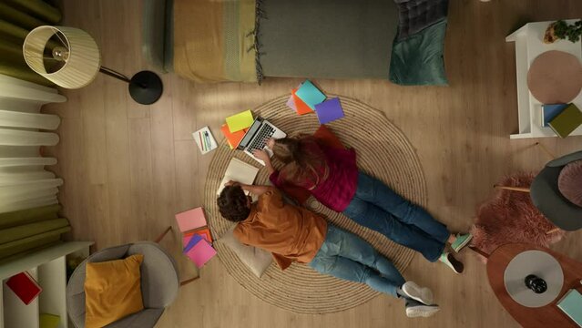 Top view capturing a young couple laying on the floor next to each other and studying, working on a laptop and taking notes, surrounded with colorful books.