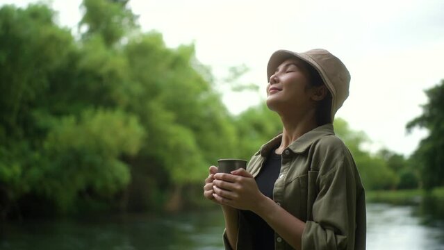 Happy woman breathing fresh air and drinking coffee near river forest campsite in the morning. Relax and vacation time with nature. Human and natural concept.