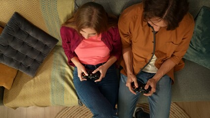 Top view capturing a young couple sitting on the couch, holding gamepads, acting as if they are playing a videogame together. The man seems to win the game.