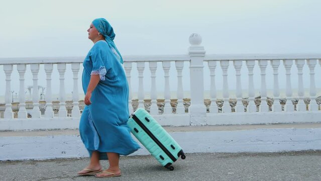 A woman in a turquoise dress and headscarf walks early in the morning with a suitcase along the embankment against the backdrop of the sea. Overall plan