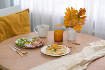 Autumn Home breakfast, vaze with yellow leaves,candle, grey plate with fried egg, sausage, bean, cup of coffee, cottage cheese with peach on round wooden table..