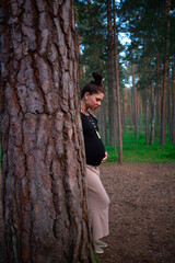 a pregnant woman stands leaning against a tree in the forest and looks at her tummy