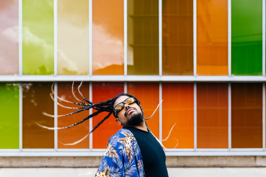 Latin man tossing his braids back in front of vibrant orange building colorful panels.