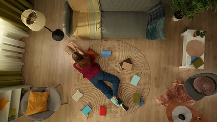 Top view capturing a young woman laying on the floor of her room, flipping pages of the book,...