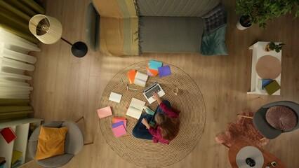 Top view capturing a young woman sitting on the floor and studying, working on a laptop and taking notes, surrounded with colorful books.