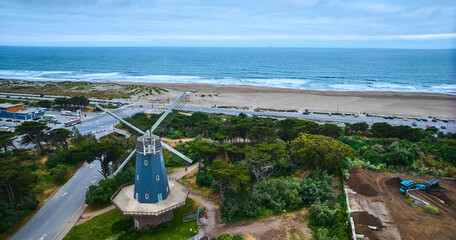 Aerial blue murphy windmill with white blades in grove of trees with sandy beach and ocean waves