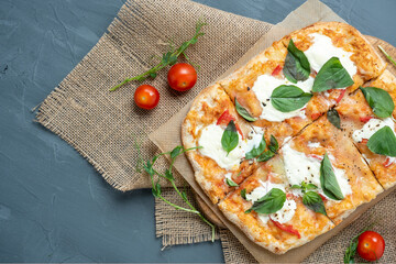 Close-up of delicious fresh crispy Roman pizza with cheese and herbs on a gray background. Fresh ripe tomatoes are nearby. Craft paper, linen napkin. Top view, flat lay