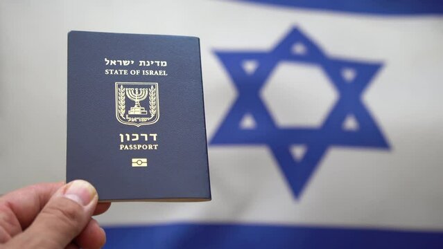 Hand of a man holds an Israeli international passport with Israeli flag out of focus waving in the wind on the background