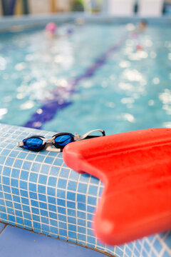 Swim goggles and kickboard at pool edge with blurred child