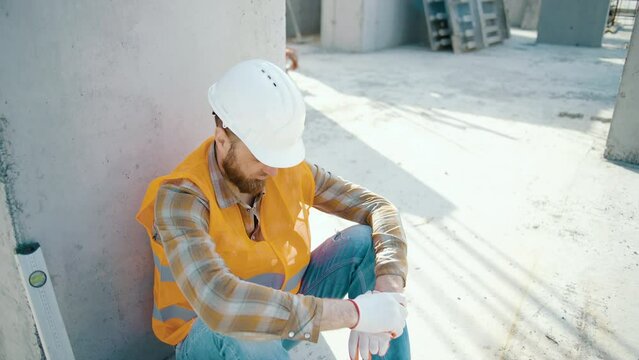Young foreman sitting outdoors and feeling tired after overwork. Caucasian worker in reflective vest and helmet at construction site