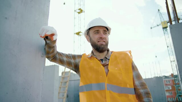 A tired worker at a construction site rests his hand on the wall after hard work and is under stress. Worker during a break at the construction of a road on a sunny summer day