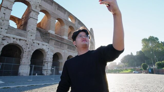 Happy young Asian tourist man selfie by mobile smartphone, Male traveller taking photo, recording vlog on holiday vacation trip at Colosseum with morning sunrise in Rome, Italy. Europe summer journey.
