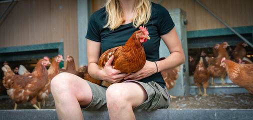 Legehennenhaltung - junge Frau sitzt an einem Hühnermobil und hält ein braunes Huhn im Arm,...