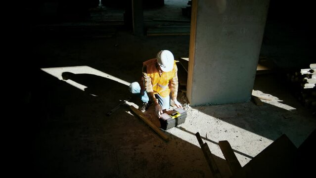 Construction worker sits near the wall and open toolbox at construction site. Shadows from man
