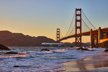 Ocean liner going under Golden Gate Bridge at sunset from sandy beach with waves