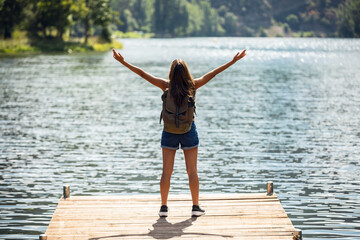 Back view of young woman relaxing and raising his arms while staying near the lake in a mountain landscape