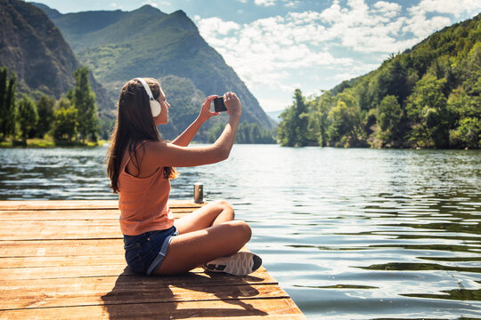 Happy woman listening to music with headphones while taking photos with smartphone to the lake