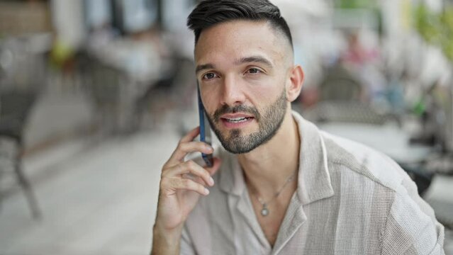 Young hispanic man talking on smartphone drinking coffee at coffee shop terrace