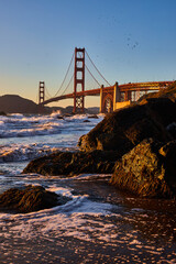 Golden water around black boulder on shore with Golden Gate Bridge in distance