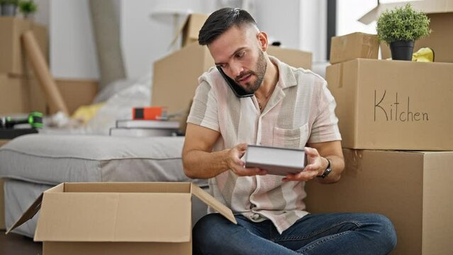 Young hispanic man packing cardboard box talking on smartphone at new home