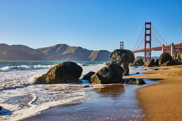 Boulders on sandy beach with waves and seafoam along shore to Golden Gate Bridge