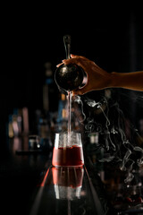 View on hand of girl bartender accurate pours steaming liquid from steel shaker into drinking glass with ice cube