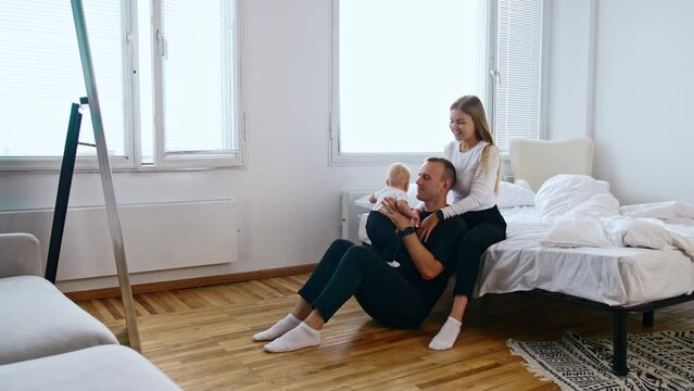 Woman sits on bed and her husband sits at her feet. Happy dad rises tiny baby above the head.