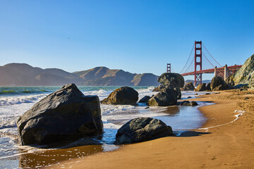 Sandy beach with waves crashing against boulders and distant Golden Gate Bridge