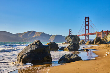 Sunny beach with waves crashing against boulders and distant Golden Gate Bridge