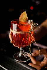 Female bartender serves glass with delicious cocktail, decorated with sprig of gypsophila and piece of peach