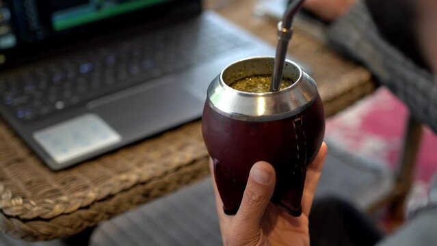 Man With His Back Turned Working On Editing A Video On A Laptop In A Hotel And Drinking Mate. Shot From Behind With Unrecognizable Person.