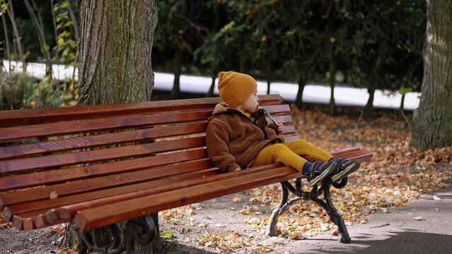 Caucasian baby sits on the bench drinking from doypack. Kid having a snack in the park in autumn.
