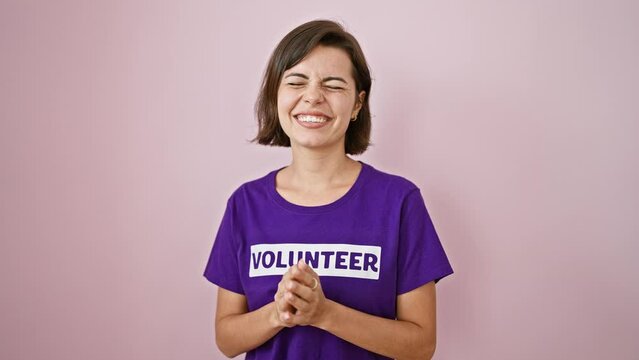 Young, confident hispanic woman celebrating her win as a volunteer, sporting a uniform t-shirt and short hairstyle, laughing in joy over an isolated pink background